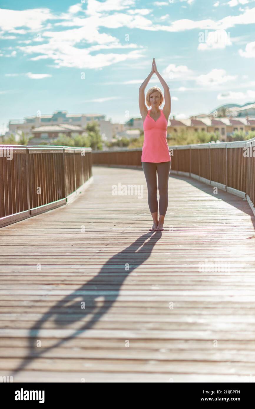 Young serious woman practicing yoga with hands raised up outdoors near on th wooden bridge against blue sky with clouds Stock Photo