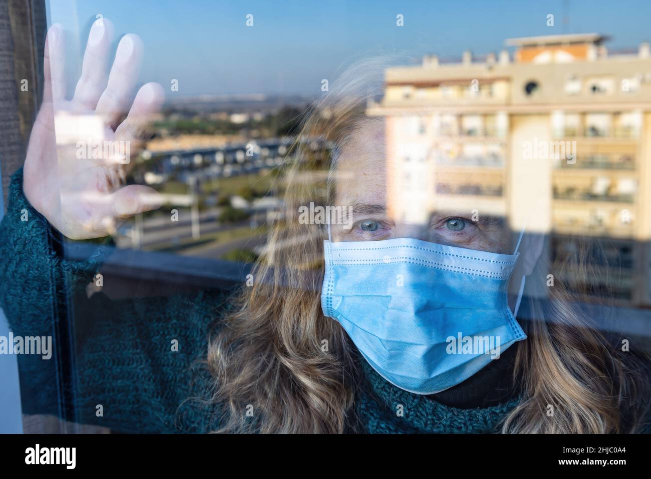 Woman isolated by coronavirus Covid-19 is looking through the glass of her house wearing a protective face mask Stock Photo