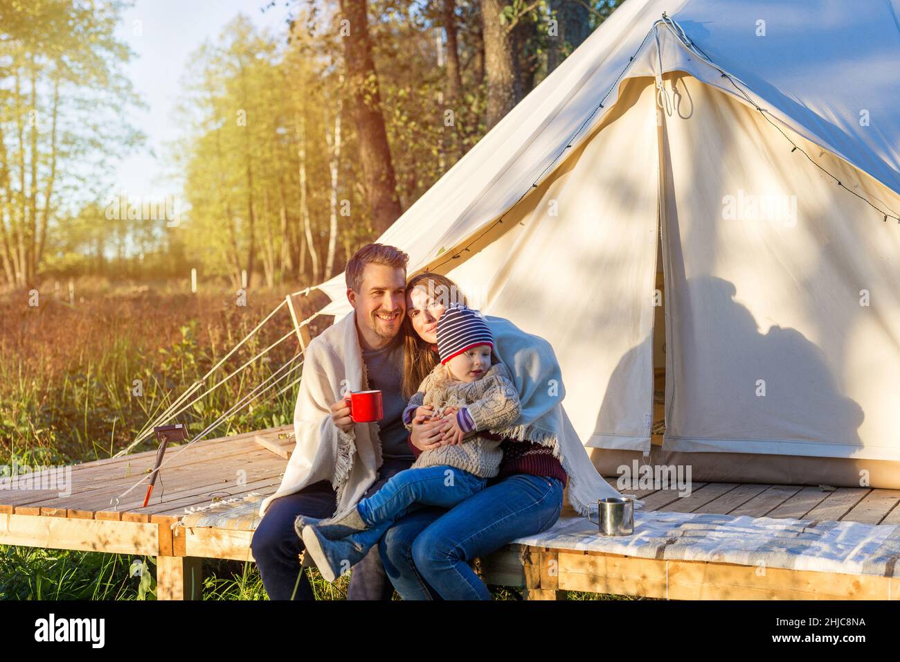 Happy young family wraps a blanket over themselves while sitting near canvas bell tent outdoors during sunset Stock Photo