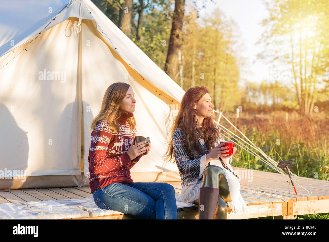 Happy women drinking some tea while sitting near canvas bell tent watching sunset Stock Photo