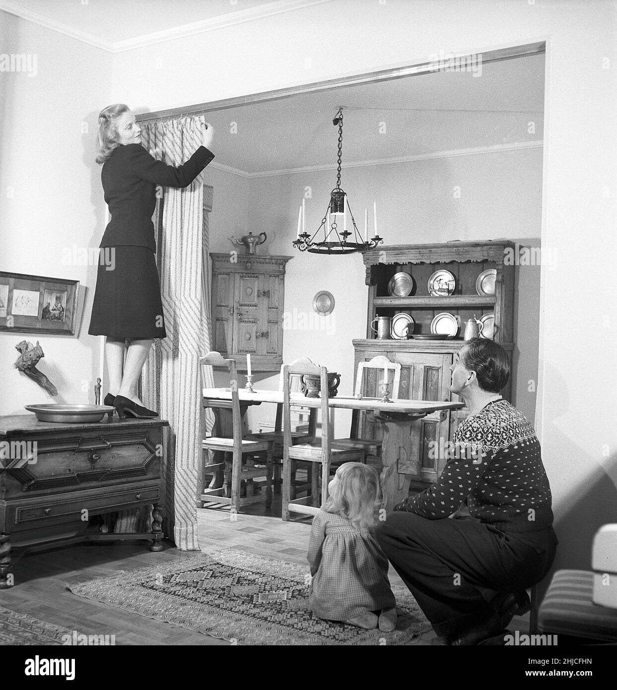 In the 1940s. A young couple arranges the drapes that divides the rooms in their apartment. Mrs Vibeke Falk stands on a piece of furniture to reach the clips and adjust the drapes while Mr Lauritz Falk and their daughter looks on. The back room has a rustic nordic interior with traditional wooden furniture with historic background. Sweden 1946 Kristoffersson ref Y38-4 Stock Photo