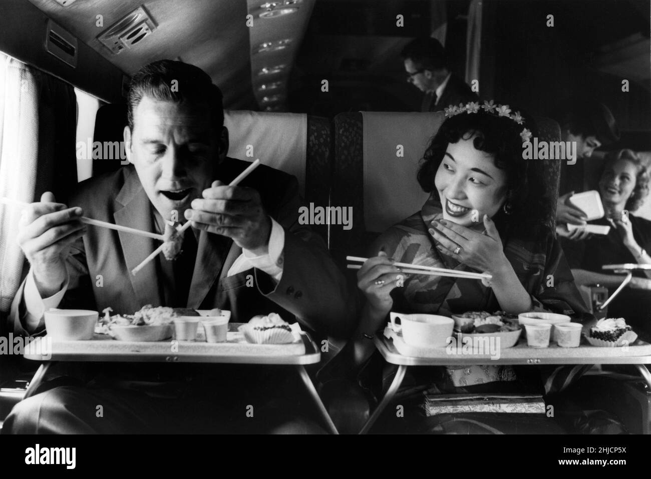 A passenger on Northwest Orient Airlines gets a lesson in using chopsticks. Circa 1960. Stock Photo