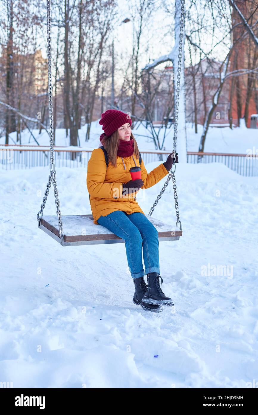 young woman swings on a huge swing in winter with a cup of coffee or a hot drink like tea. Vertical frame Stock Photo