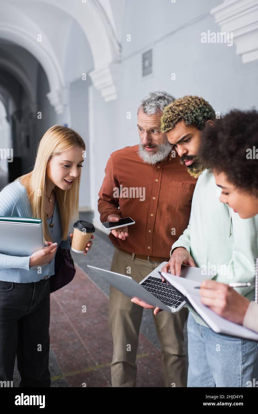 Teacher holding smartphone and talking to multiethnic students with laptops and notebooks in university Stock Photo