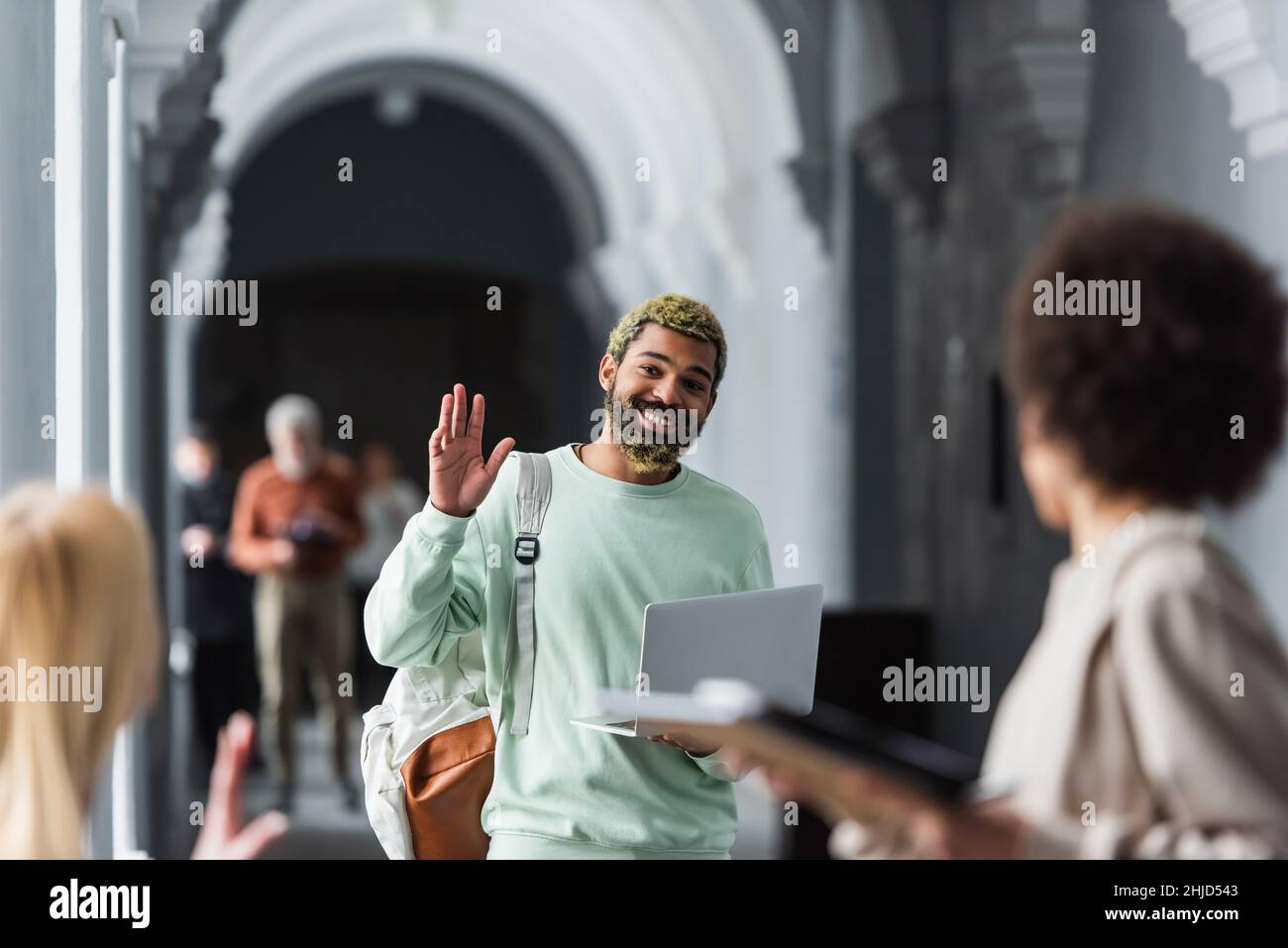 African american student with laptop waving hand at blurred friends in ...