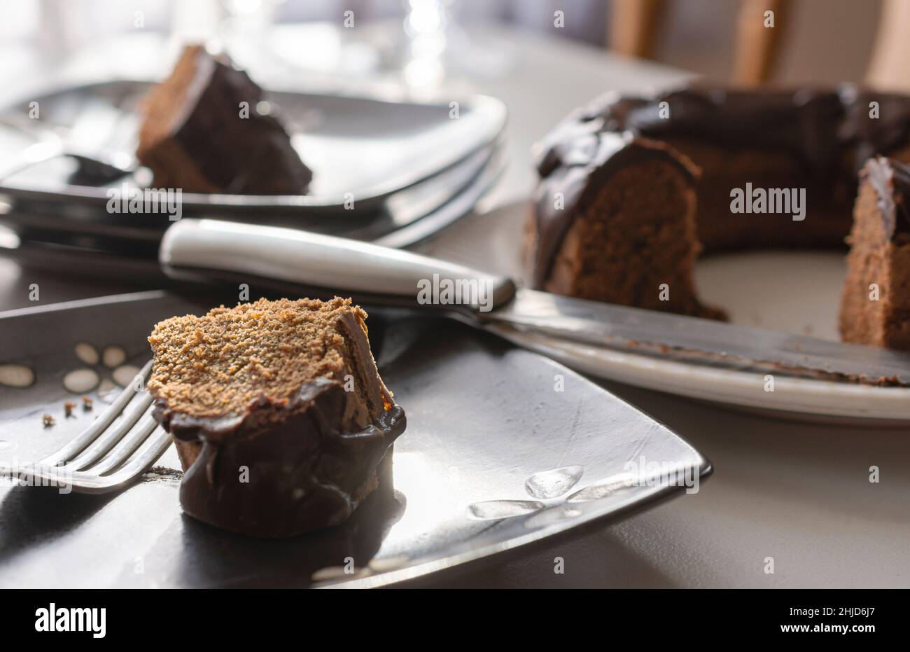 A slice of a chocolate bundt cake on a black plate with a fork.  Remainder of cake blurred in background along with an additional slice of cake on a s Stock Photo