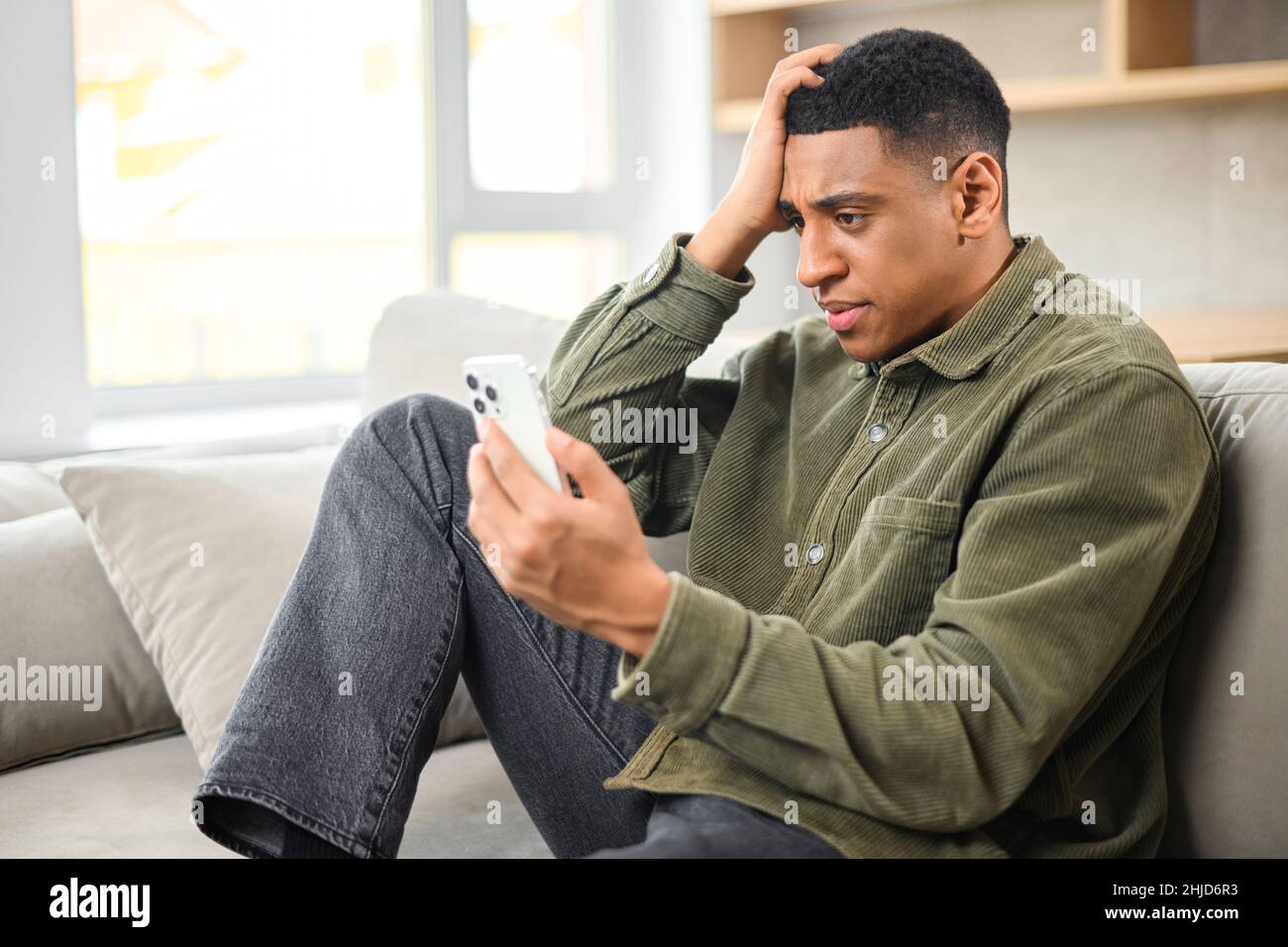 Sad young multiracial man holding smartphone and sitting on the sofa at modern apartment while looking with shocked face and holding his head Stock Photo