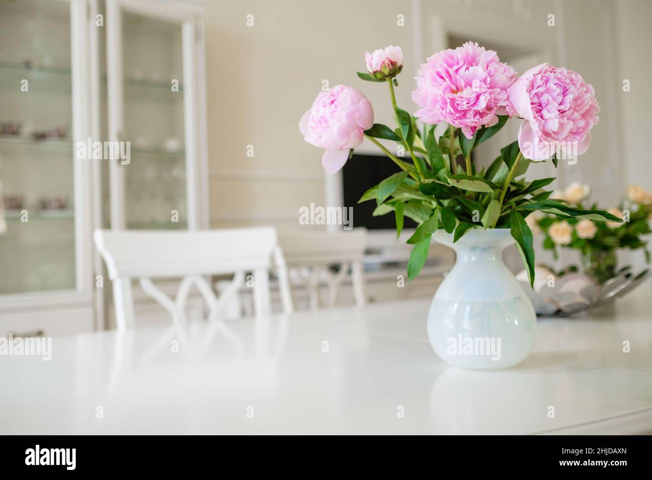 Pink blooming peony flowers with lush green leaves put in vase on white table in light room with modern furniture at home closeup Stock Photo