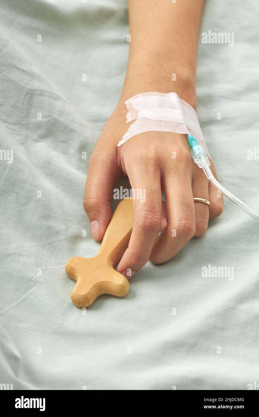 A vertical shot of a patient's hand connected to an IV drip holding a wooden cross Stock Photo