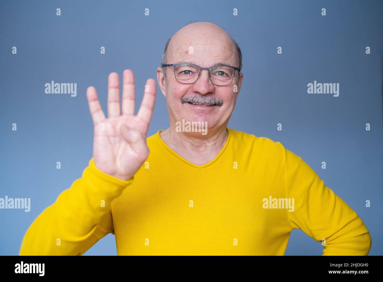 Senior man raising hand, showing number four with fingers, counting something, standing over blue background Stock Photo