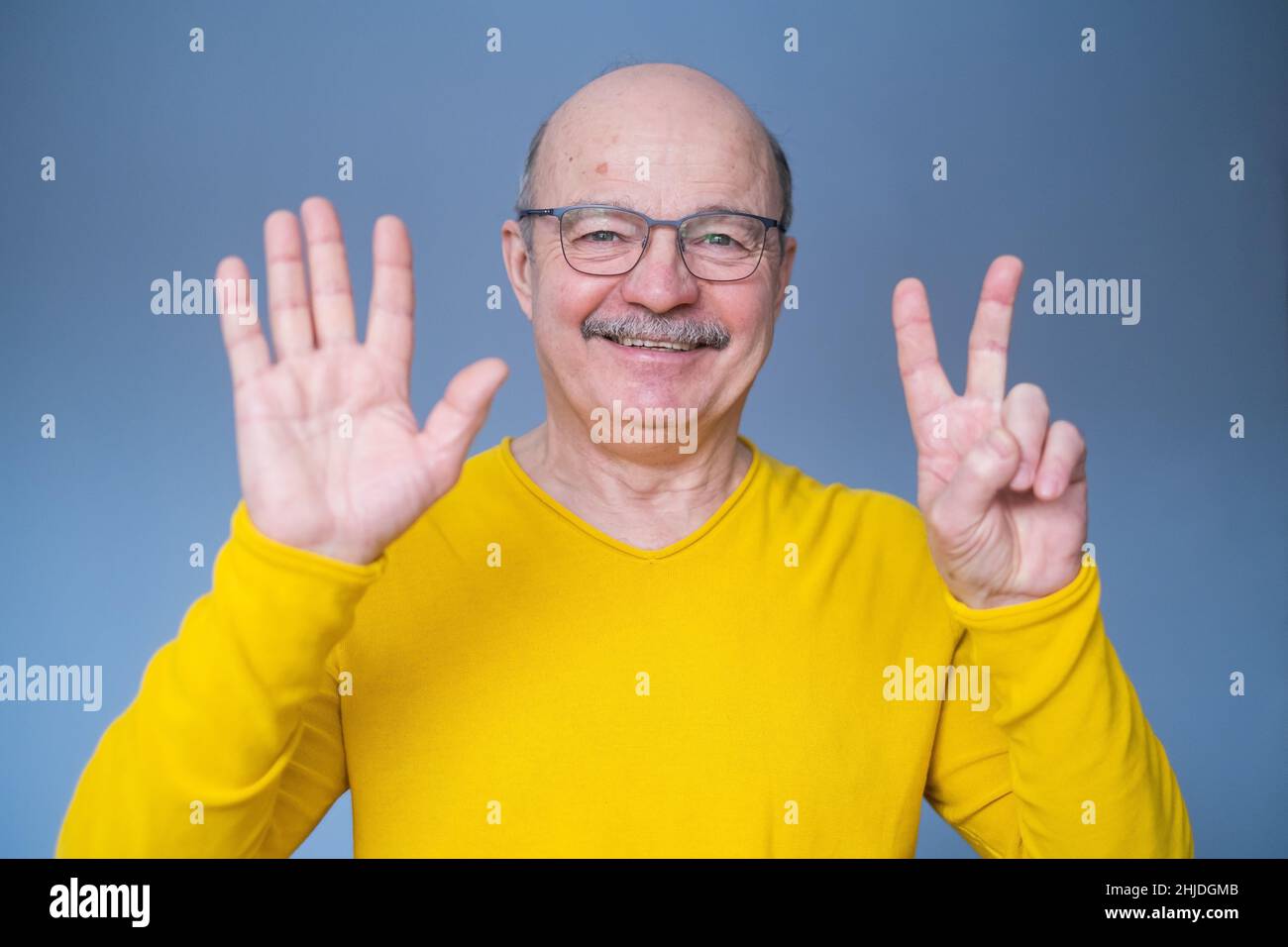 Senior man raising hand, showing number seven with fingers, counting something, standing over blue background Stock Photo