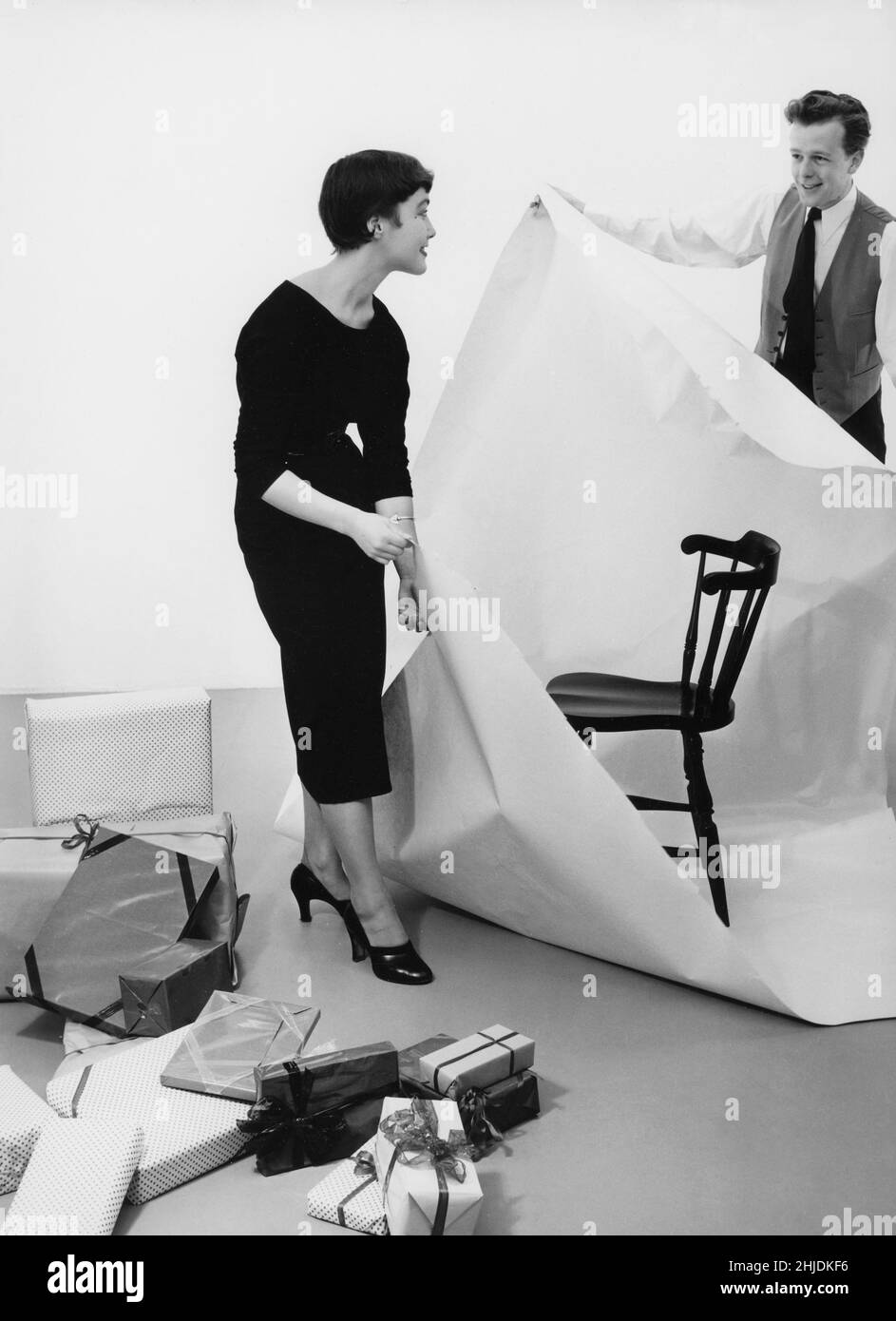 Christmas in the 1950s. A couple with christmas gifts visible tries their best at wrapping a chair in paper. 1953 Stock Photo