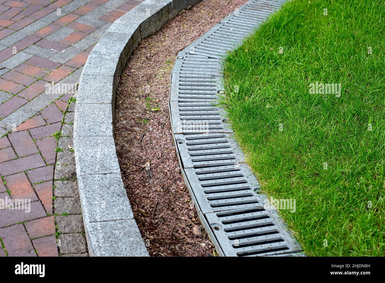 iron drainage grate on the roadside with green grass and stone pebbles at the granite curb along the pedestrian pavement of brick tiles, nobody. Stock Photo