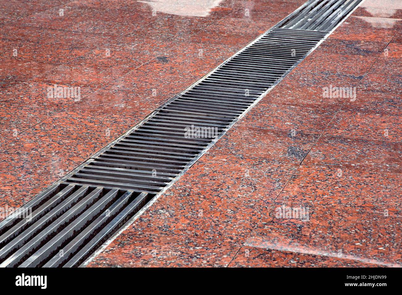 drainage grate with stripes on the floor with a wet red stone granite surface, close-up of a storm system wet after rain. Stock Photo