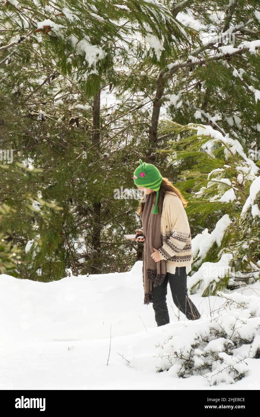 Mevasseret Zion, Israel - January 27th, 2022: A teenage girl walking in a snowy pine forest, looking at her smart phone. Stock Photo