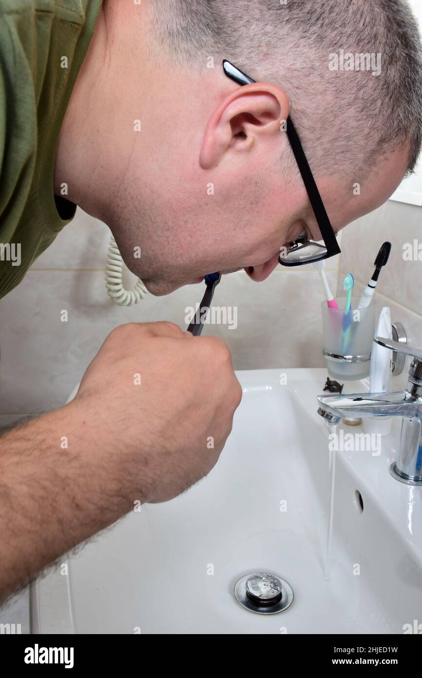 A white man with glasses is brushing his teeth in a bathtub above the sink. He doesn't look at the camera. The tap is open and water is flowing. Stock Photo