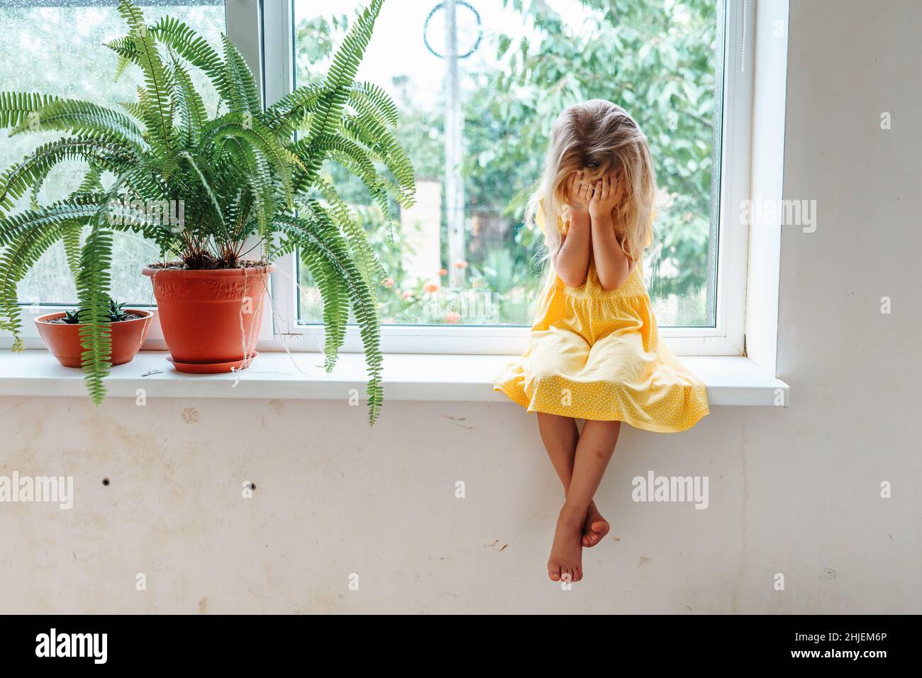 Young little crying girl barefoot sitting on window sill face in hands near plants. Desperate child. Side view.  Stock Photo