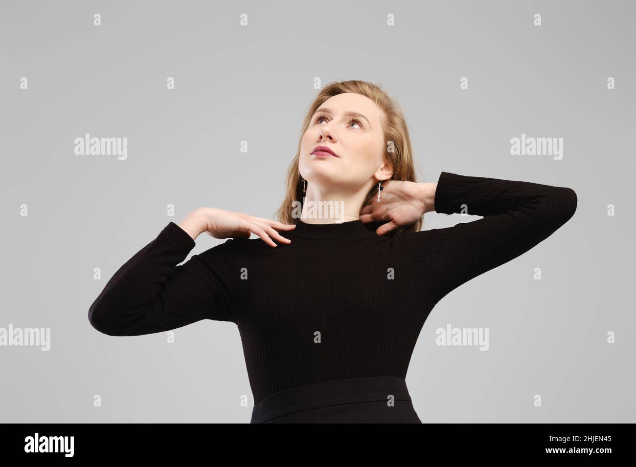 Low angle photo of young woman lifting hands to her shoulders and looking up in studio over grey background Stock Photo