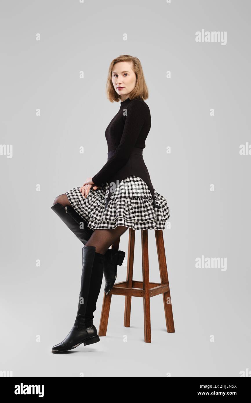 Young woman in turtleneck and suspender belt over little skirt sitting on tall chair in studio Stock Photo