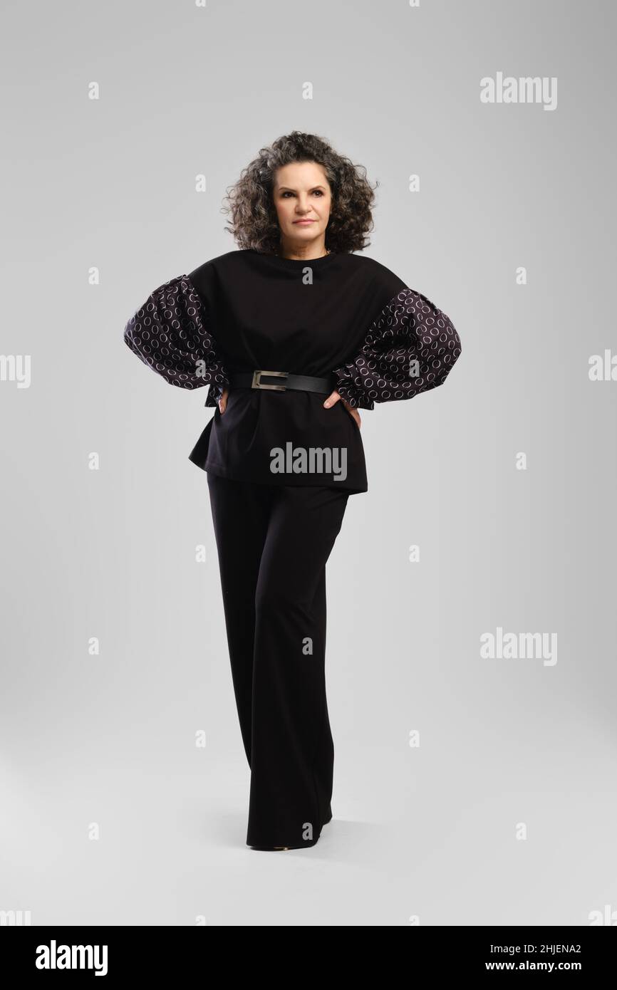 Senior woman with lush curly hair and black pantsuit posing in studio with hands on waist over grey background Stock Photo