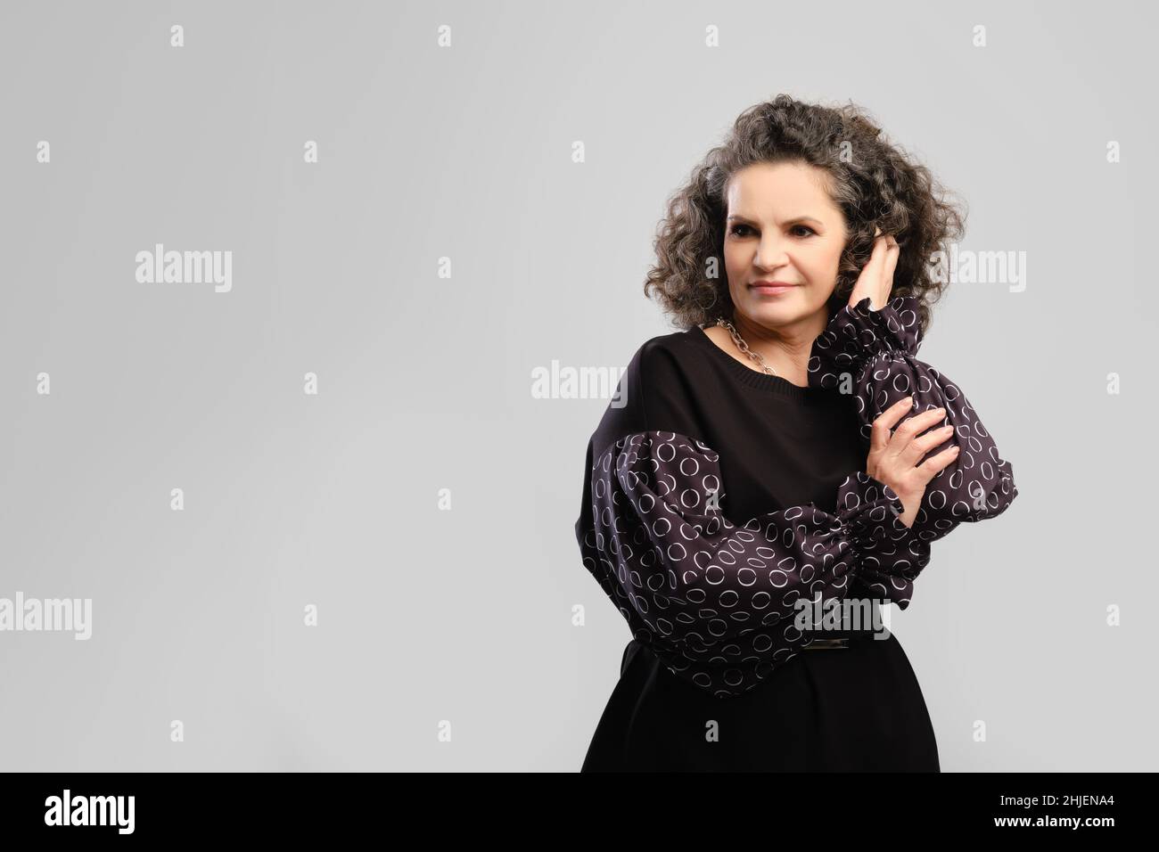 Senior woman touching lush curly hair in studio over grey background Stock Photo