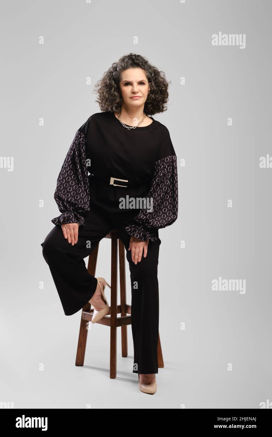 Senior woman with lush curly hair and black pantsuit sitting on tall chair in studioo over grey background Stock Photo