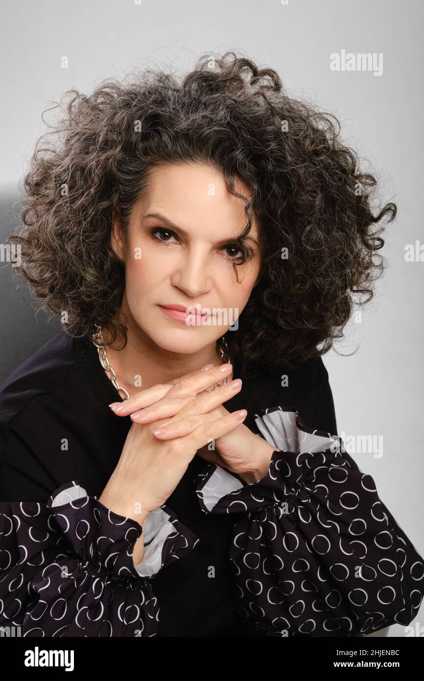 Closeup portrait of senior woman with lush curly hair and black pantsuit posing in studio over grey background Stock Photo