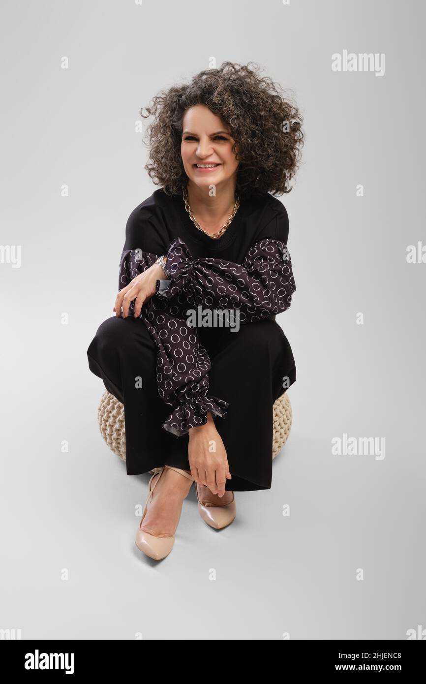 Happy senior woman with curly hair sitting on padded stool in studio over grey background Stock Photo