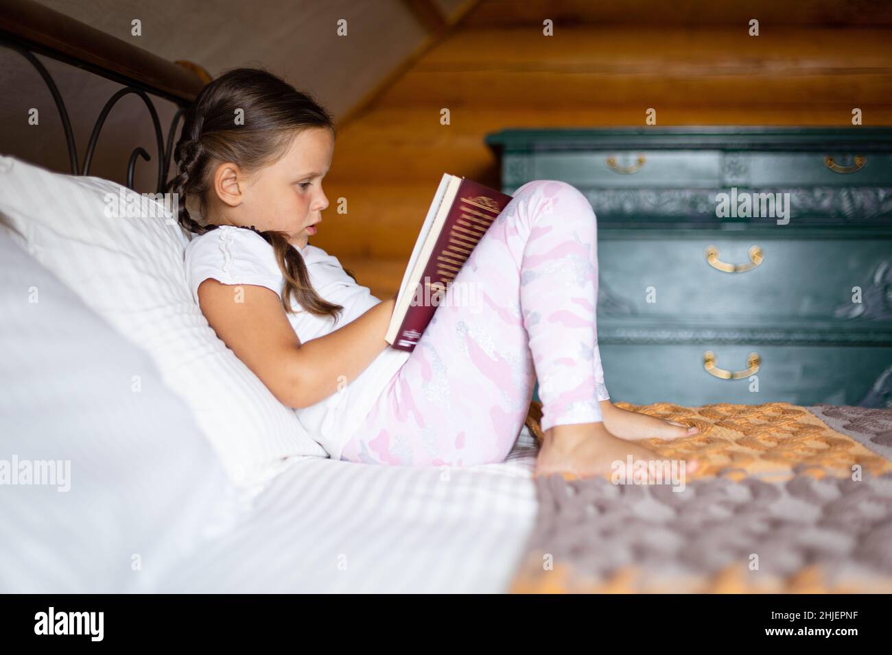 Caucasian keen child having book in hands reading with great attention lying on bed wearing pajamas in wooden house. Free time leisure activities Stock Photo