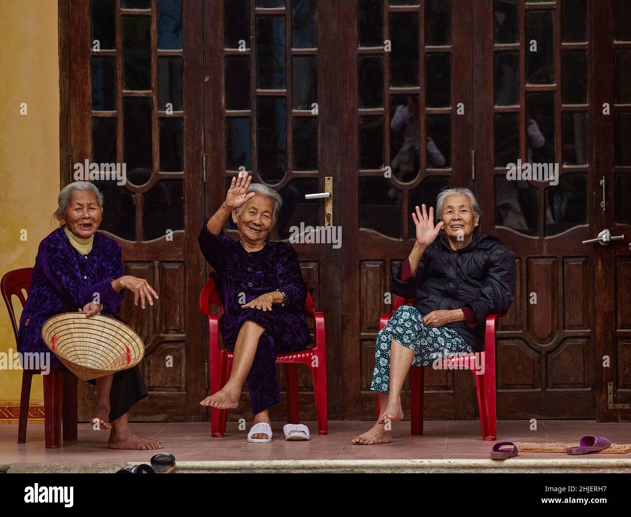 Three elderly Vietnamese women sit on chairs in front of the house. The women smile and greet with a raised hands sincerely. Wood carving doors. Stock Photo