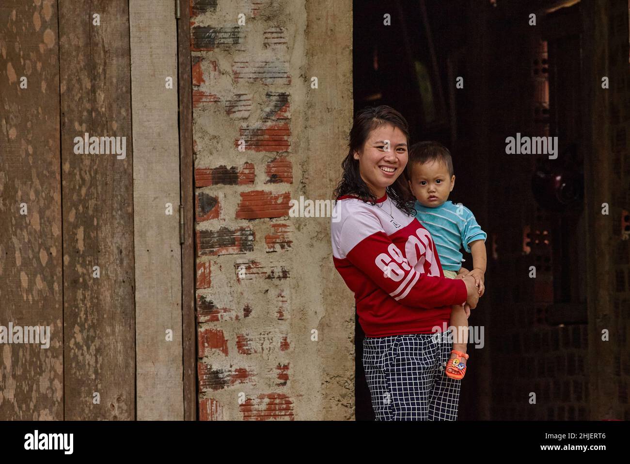 Young smiling Vietnamese woman holds her child on the hands in front of a dark house entrance. Stock Photo