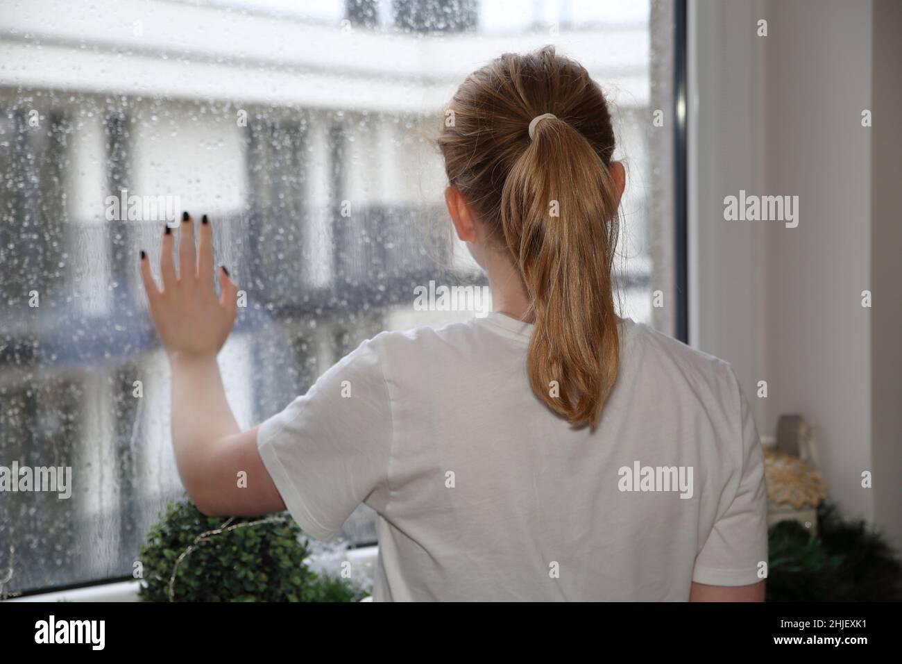 Blond-haired girl stand at the window touching glass by her hand, lonely, behind urban area, rainy deppresive weather Stock Photo
