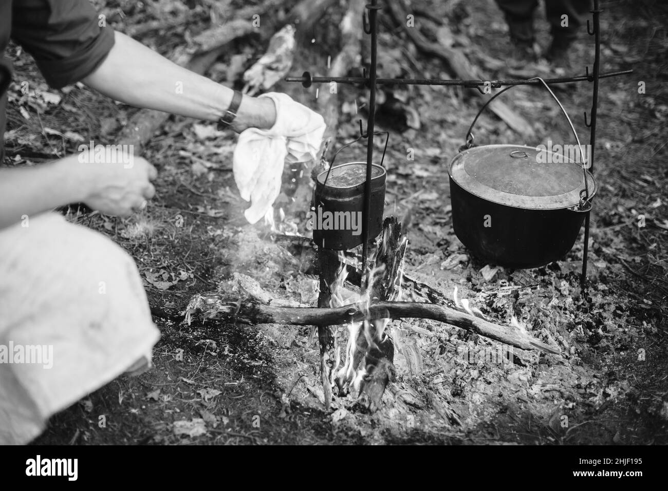 Re-enactor Dressed As World War II German Wehrmacht Soldier Cooking Food Over A Fire In An Old Marching Pot. Photo In Black And White Colors. Soldier Stock Photo
