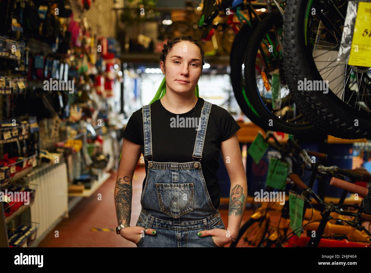 Unemotional young female seller with dreadlocks and tattoos on arms looking at camera while standing in store with hands in pockets of denim overall Stock Photo