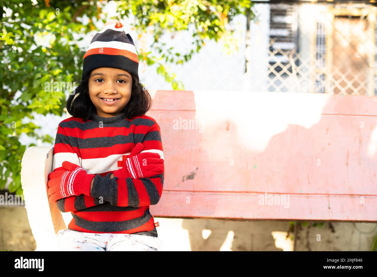 Indian smiling girl kid with tight crossed arms due cold sitting at park by looking at camera - concept showing of holidays, winter wear with copy Stock Photo