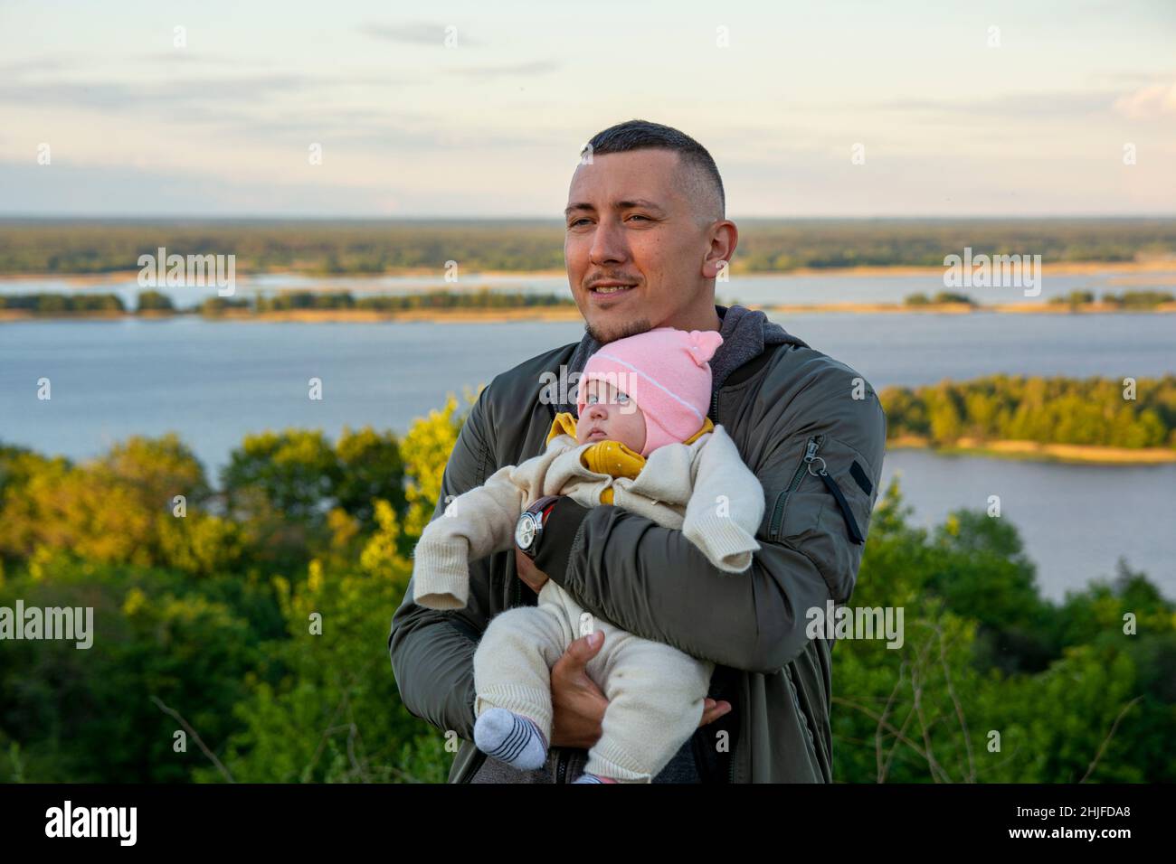 A young father with a tiny baby in his arms. The father loves his child. Photos of dad with a child against the backdrop of a large river, sunset sky Stock Photo