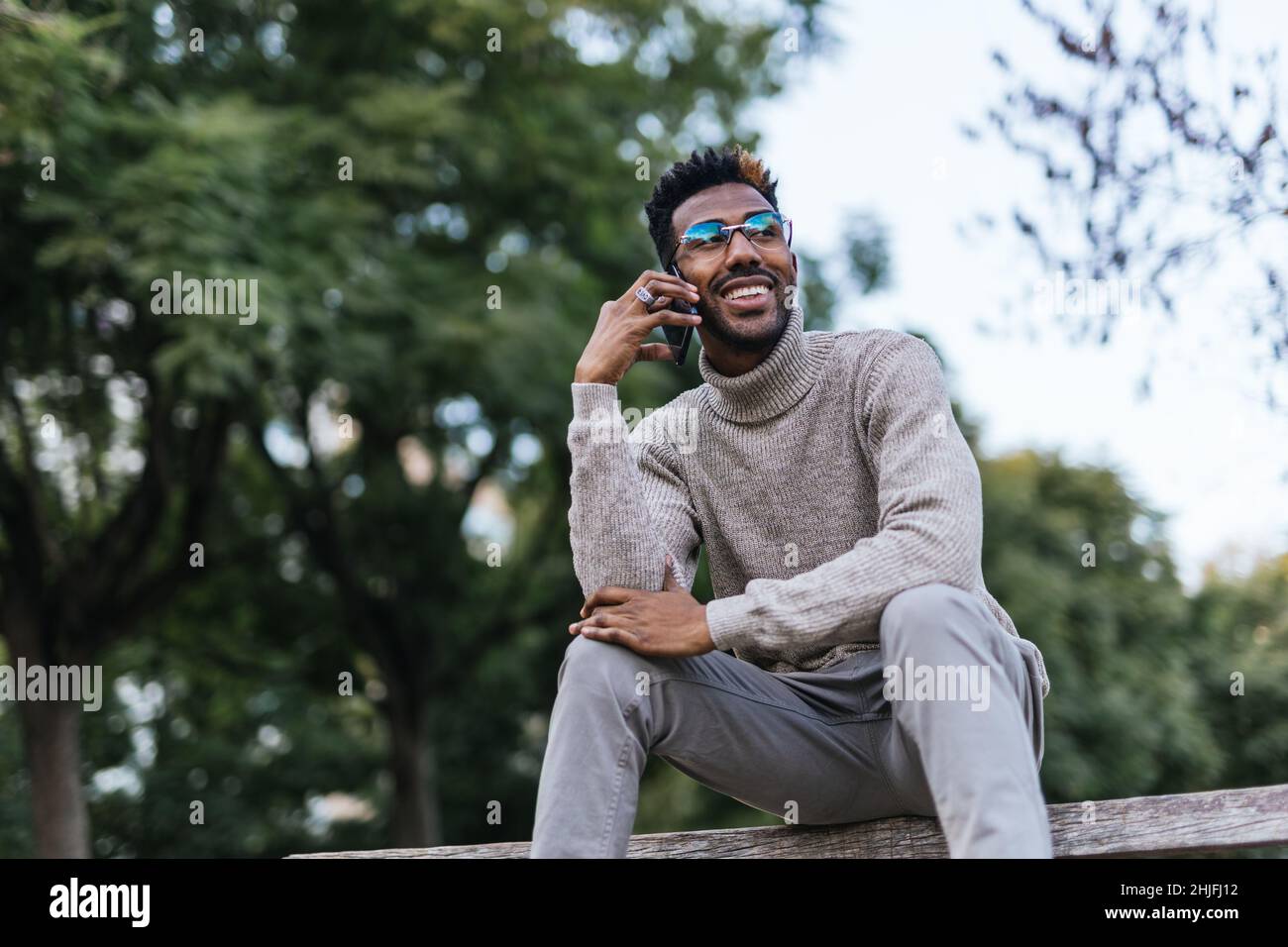 Smiling African American man in turtleneck vest sitting on a park bench and talking on his mobile phone Stock Photo