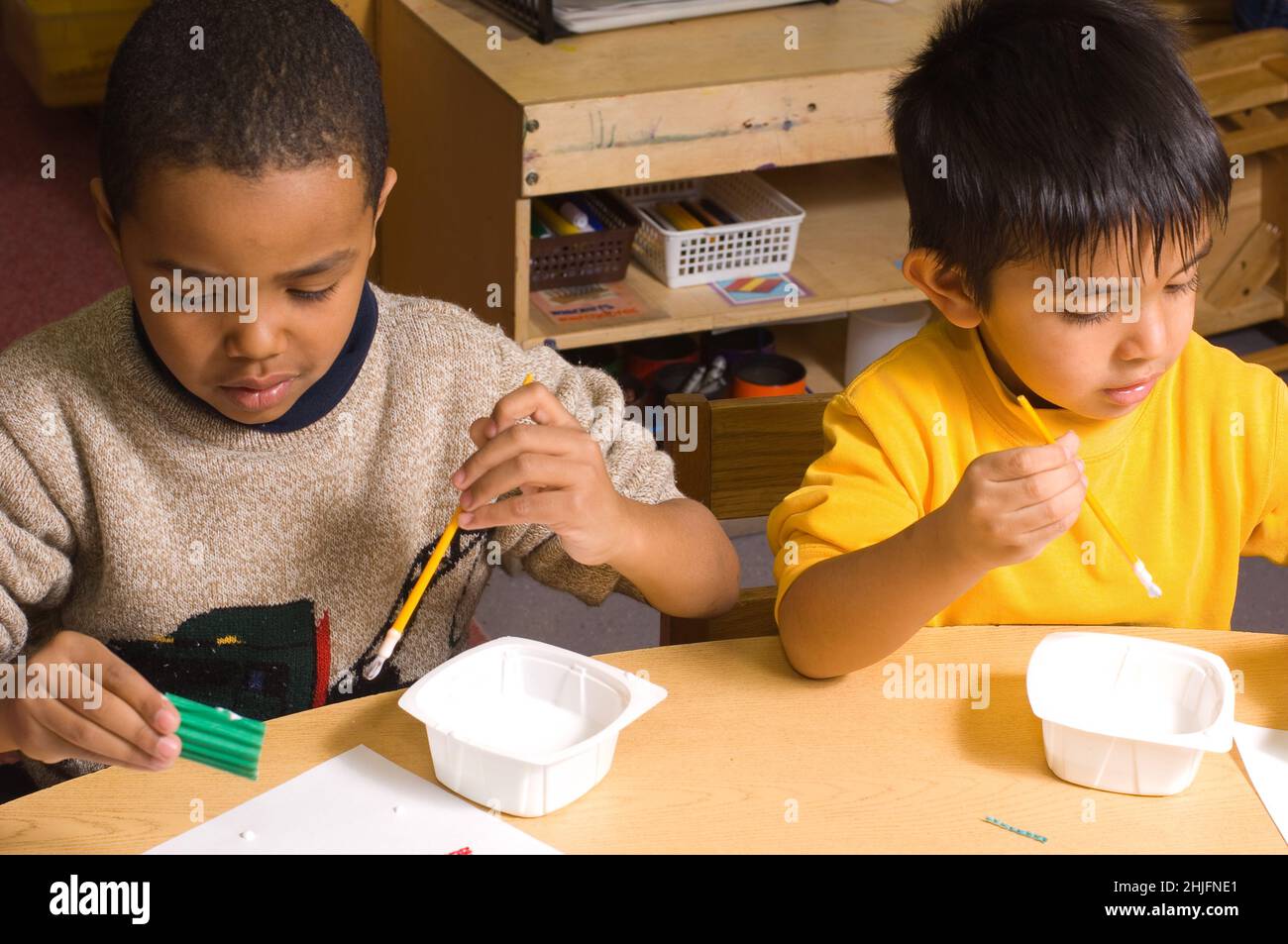 Education Preschool classroom 4-5 year olds art activity two boys sitting side by side using paint brushes to apply glue, using opposite hands Stock Photo