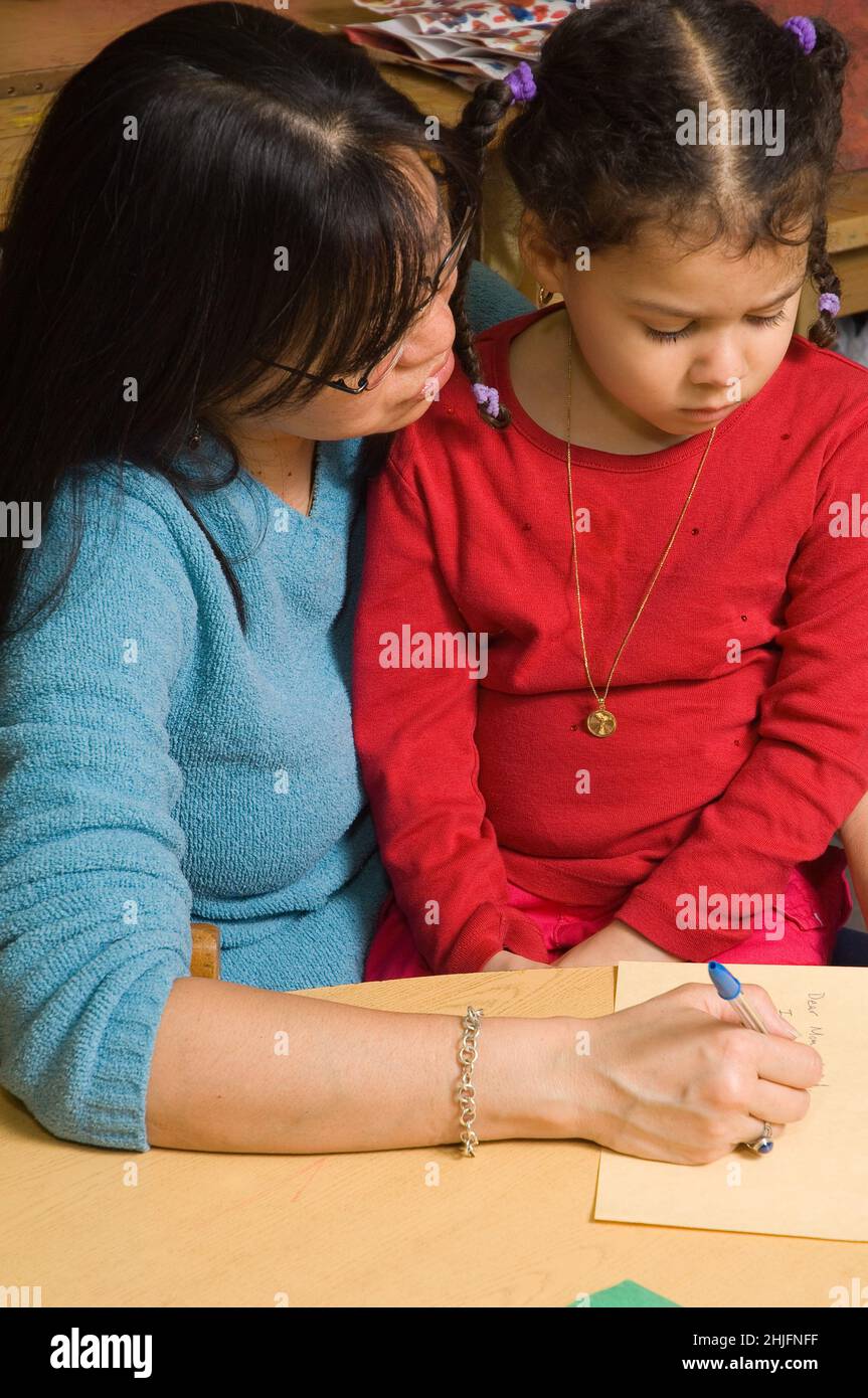 Education Preschool classroom 4-5 year olds female teacher with girl writing down what the girl wants to say about her drawing Stock Photo