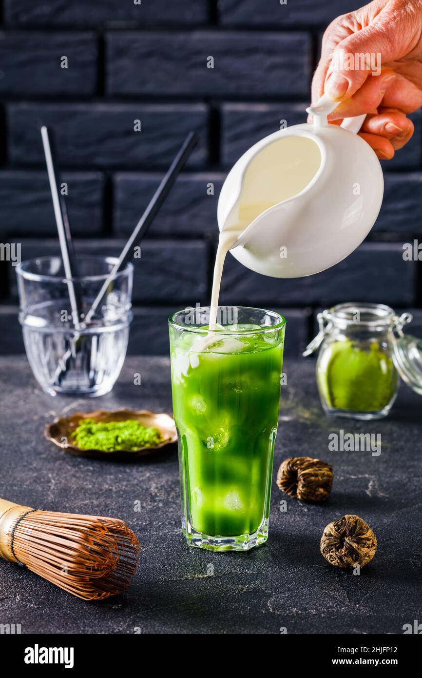 barista hand pouring milk in a glass with Iced Green matcha tea or match latte mixed with ice cubes with a brick wall at the background and powdered m Stock Photo