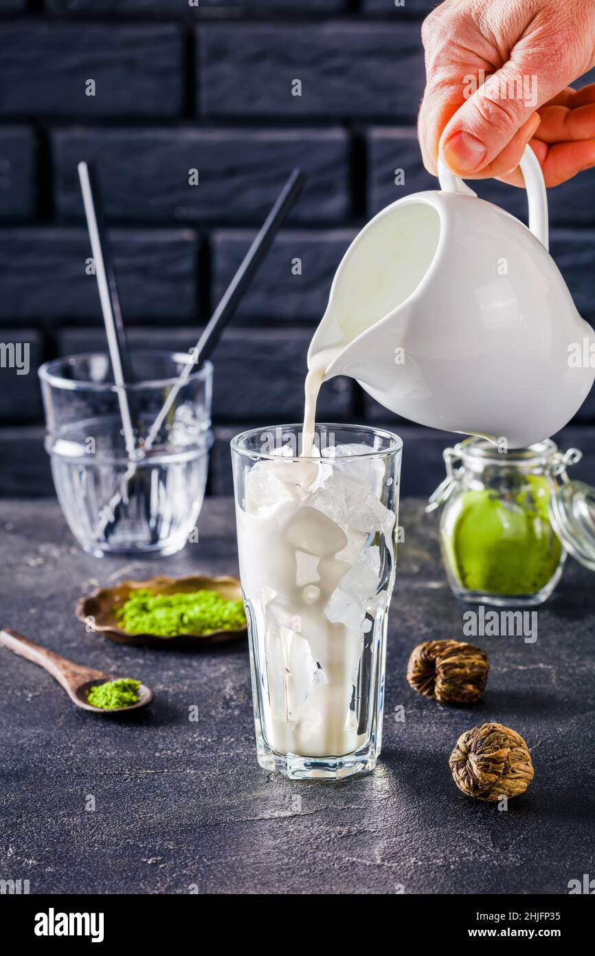 barista hand pouring milk in a glass with ice cubes with a brick wall at the background, powdered matcha tea in glass jar, close up, vertical view Stock Photo