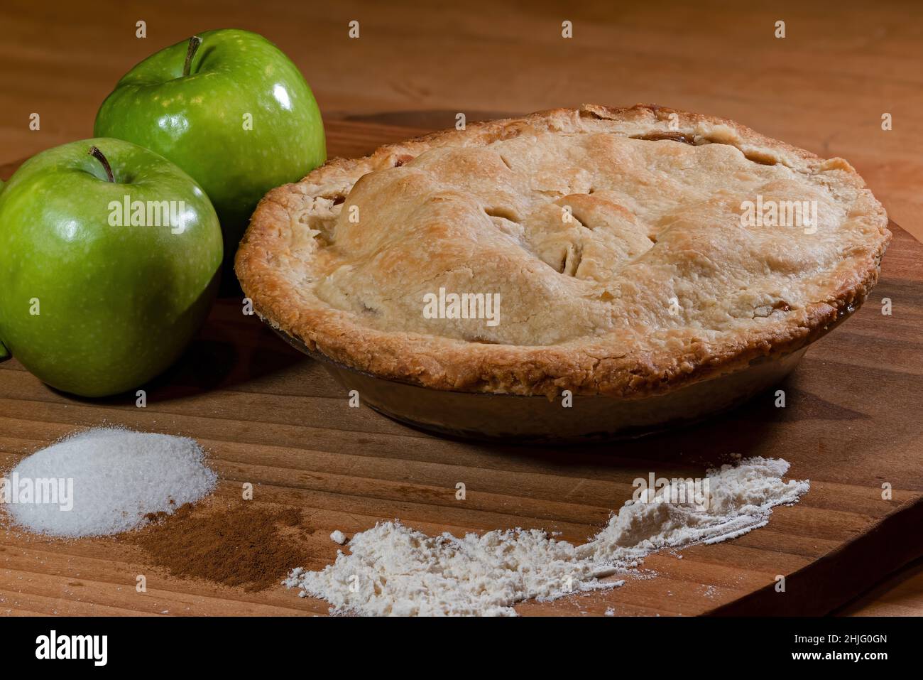 Fresh baked double crust apple pie displayed with key ingredients of green tart apples, sugar, cinnamon, and flour. Stock Photo