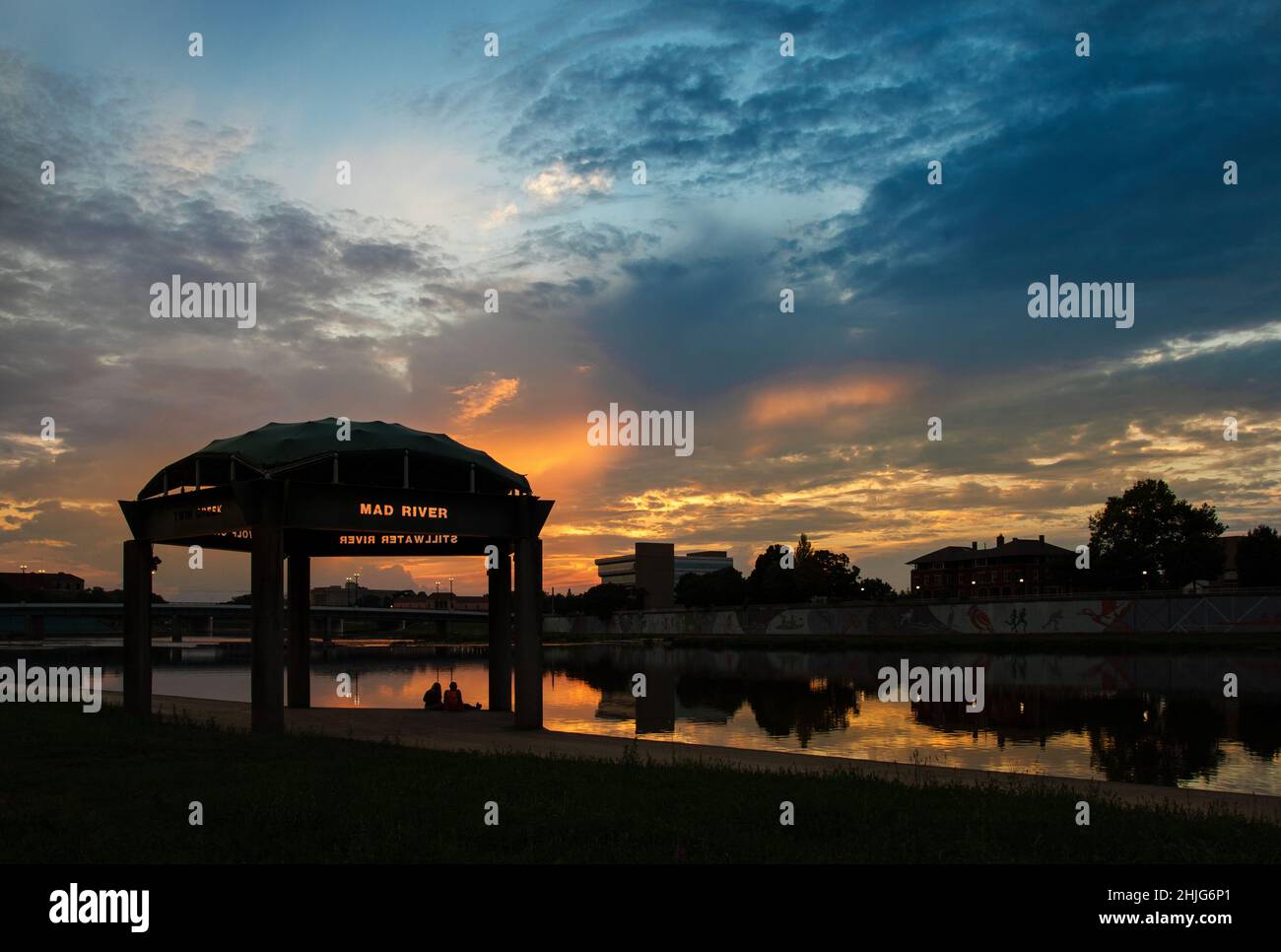 Landmark pavilion, with two people seated, at the confluence of the Mad River and Miami River Riverscape. Sunset sky is in the background. Riverscape Stock Photo