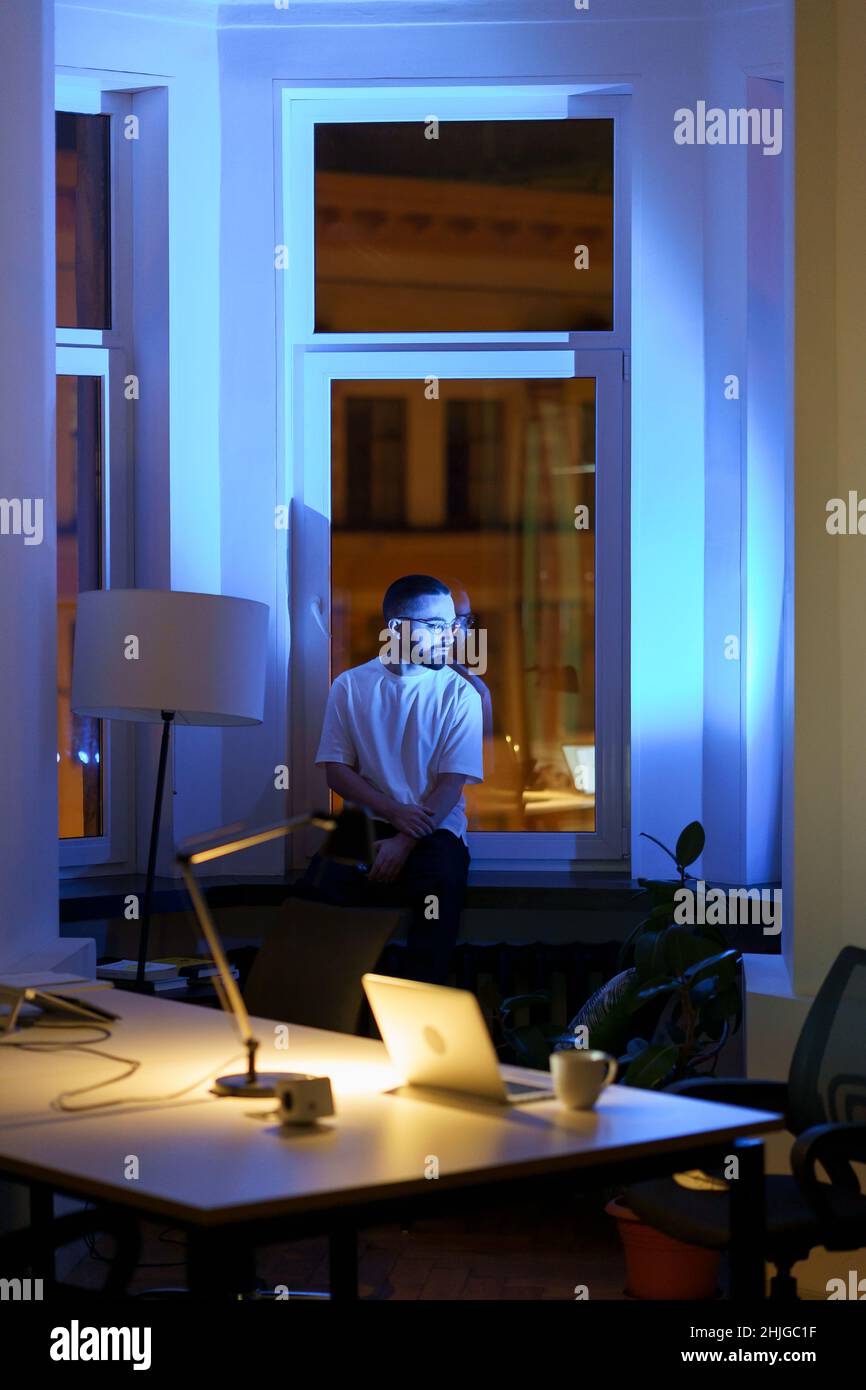 Pensive manager or business owner sit on window sill in night office thinking on problem solution Stock Photo