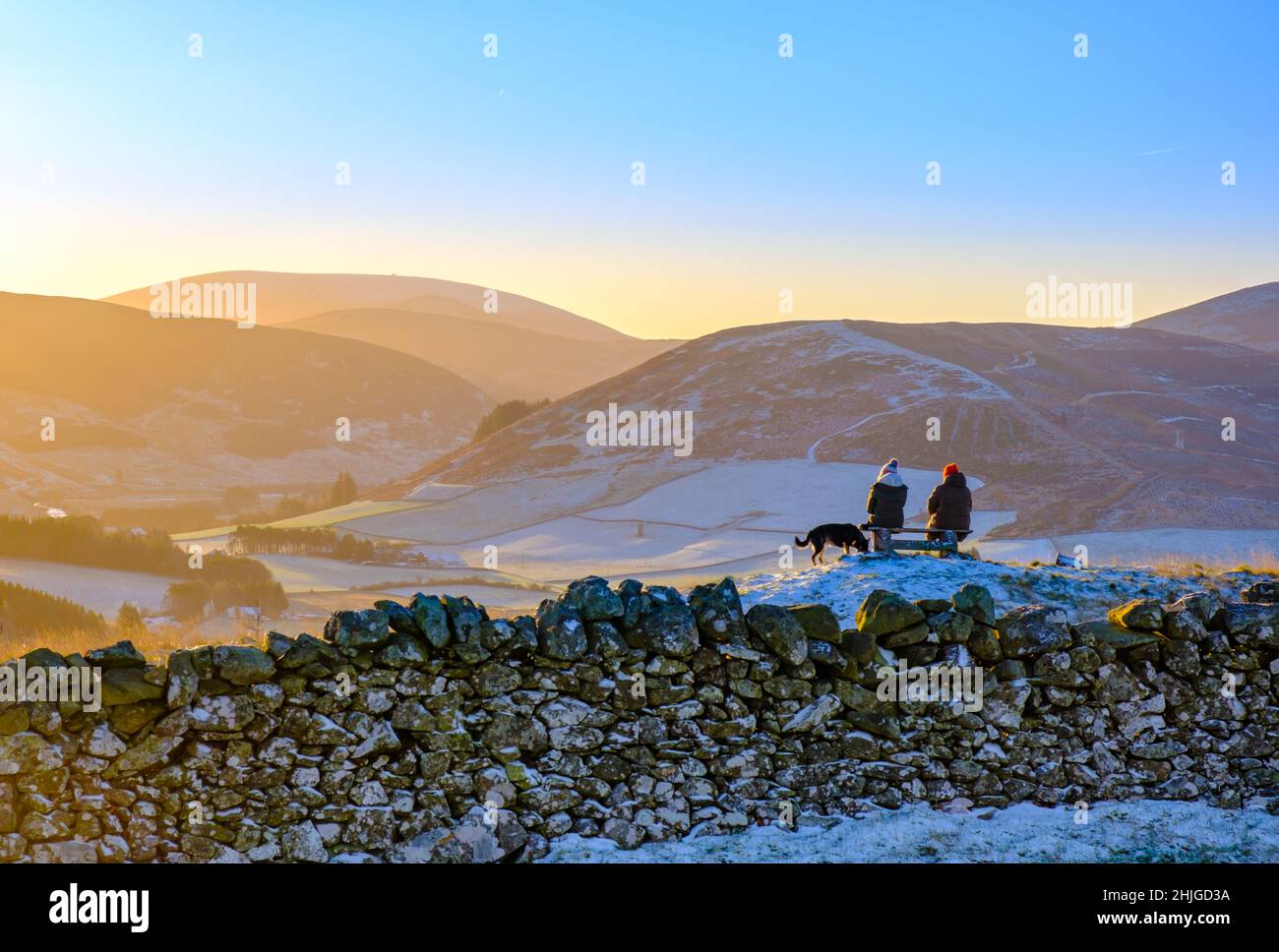 Two Friends Sitting On A Bench In The Beautiful Scottish Countryside After A Hike On A Frosty Morning, With Their Dog Stock Photo