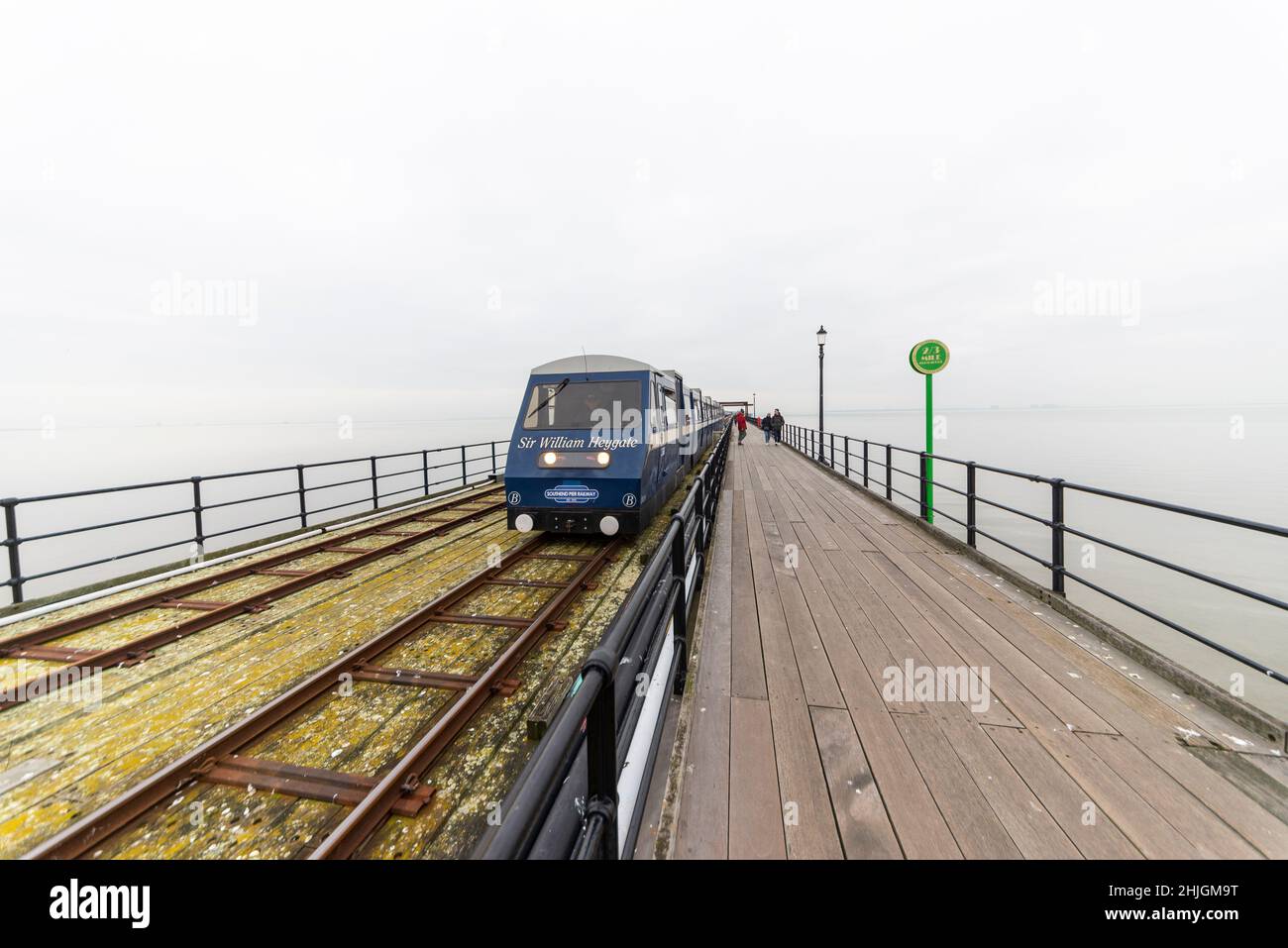 Southend Pier Railway train coming out of a misty white Thames Estuary on a still winter morning. Few people walking on walkway. Distance marker Stock Photo