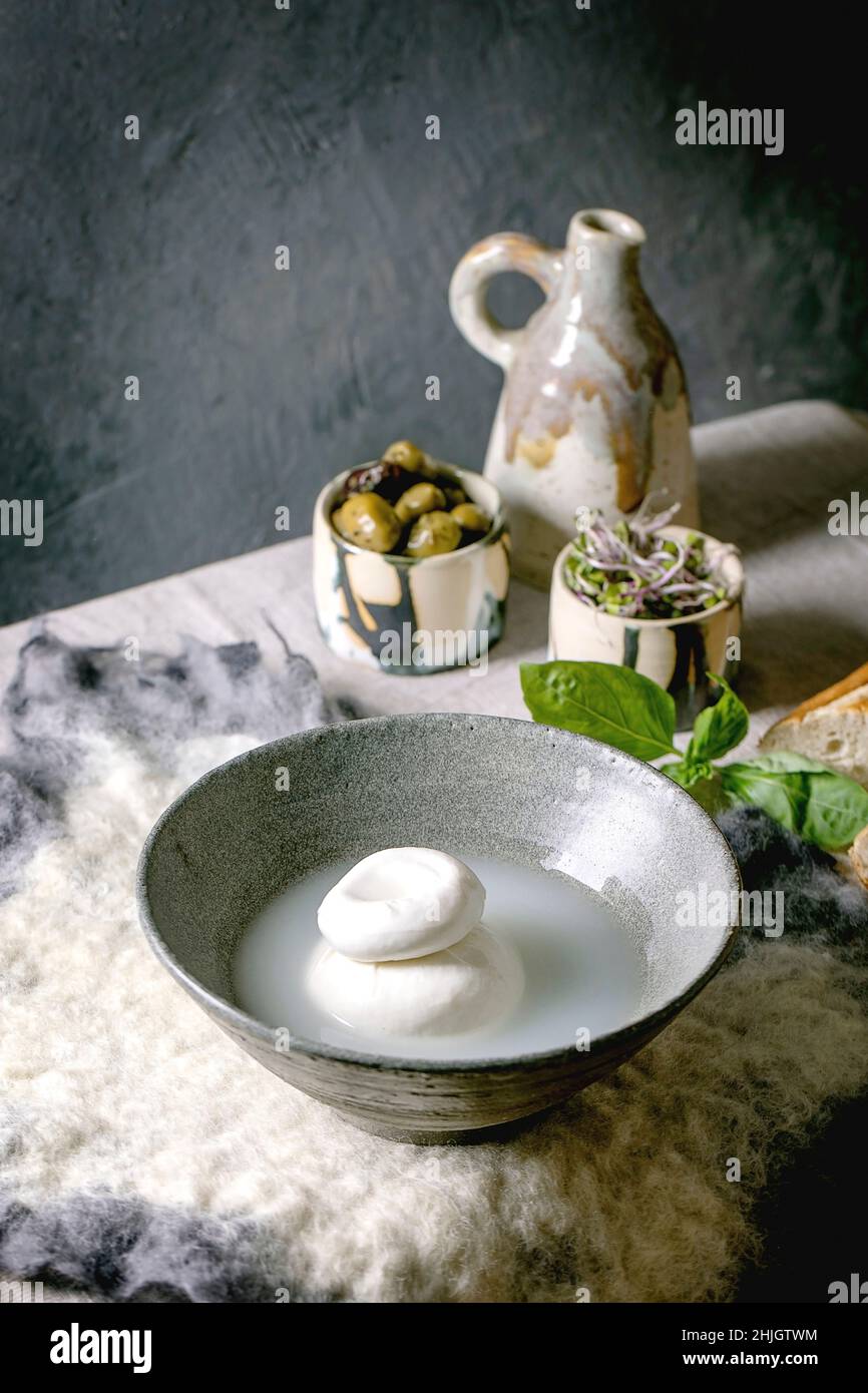 Traditional italian burrata knotted cheese in brine in grey ceramic bowl on table. Bread, olives, green salad around. Ingredients for healthy mediterr Stock Photo