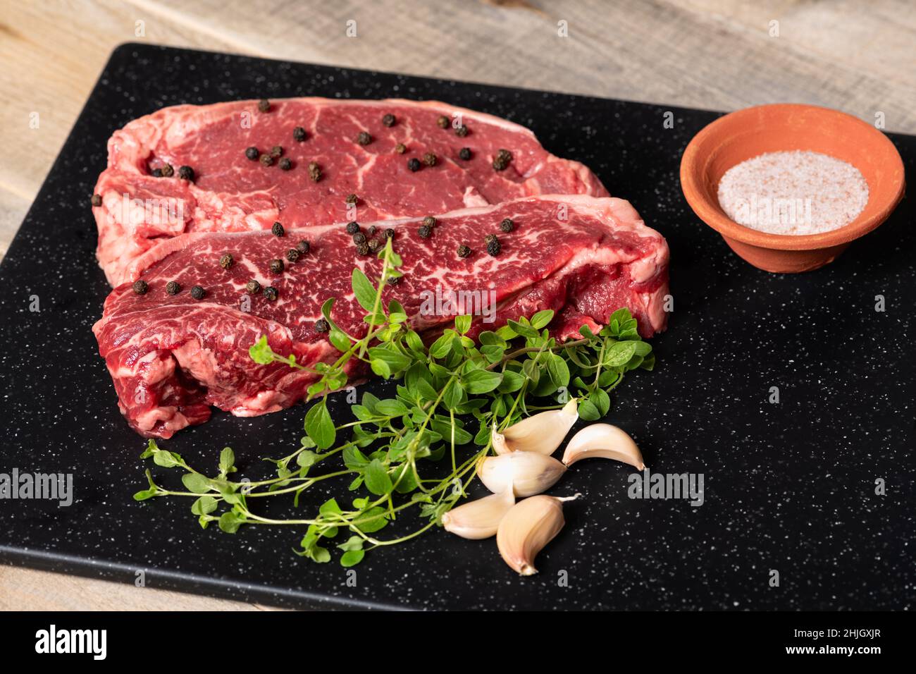 Two raw marbled beef steaks, ready for cooking, with fresh oregano, garlic, and peppercorns. Stock Photo