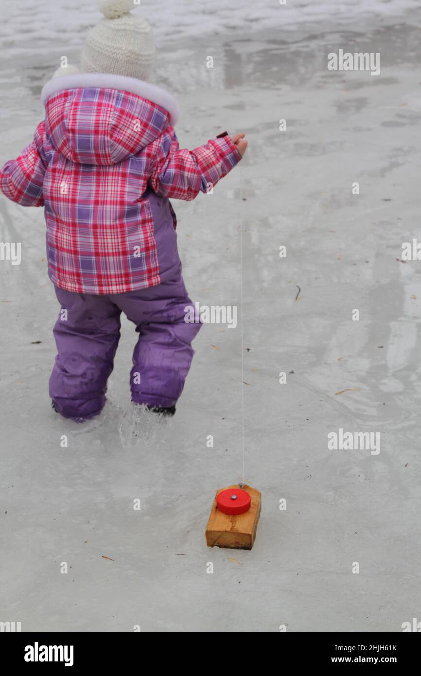 Girl in snowsuit pulling a homemade wood boat on melting ice Stock Photo