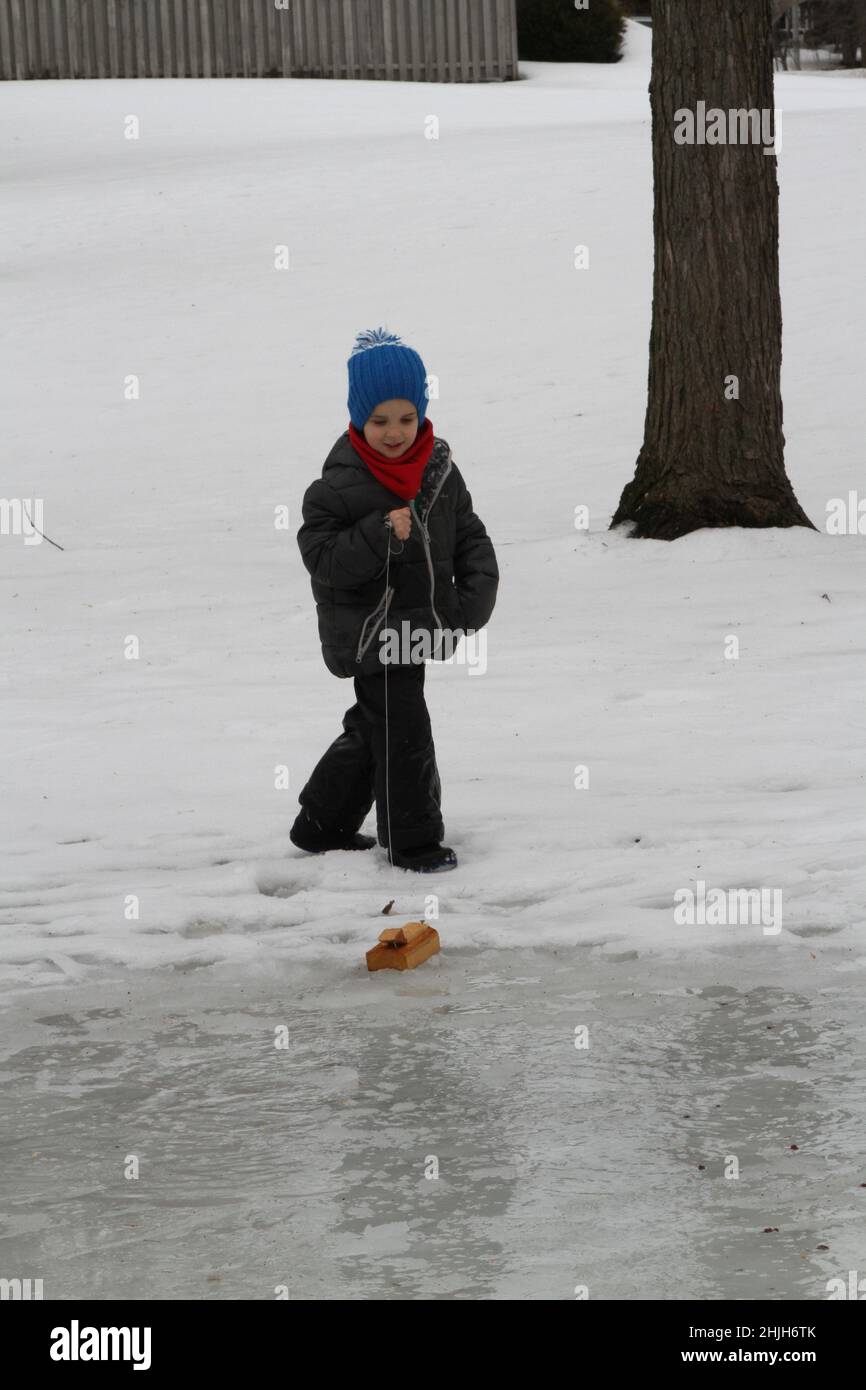 Young boy in snowsuit pulling a handmade boat on wet ice Stock Photo